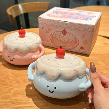 Two cute ceramic containers shaped like cakes with a hand holding one, on a wooden table with a box in the background.
