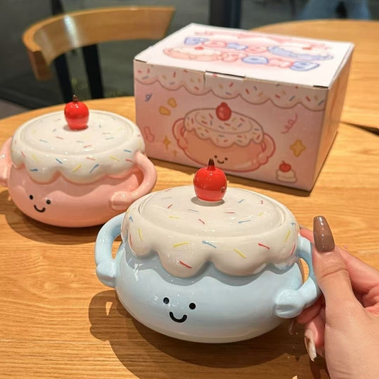 Two cute ceramic containers shaped like cakes with a hand holding one, on a wooden table with a box in the background.