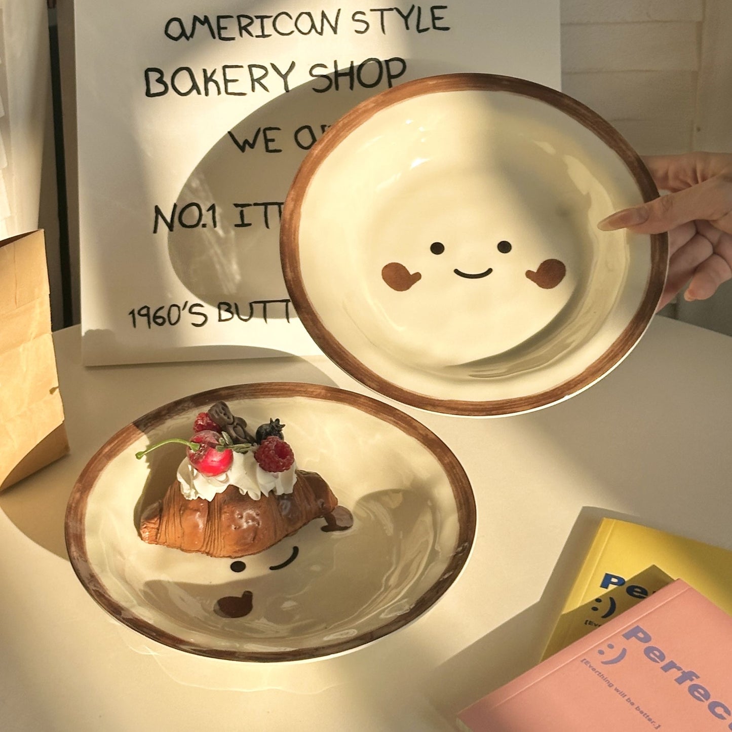 An 8-inch cream ceramic plate featuring a hand-painted brown smiley face and rim, pictured with a chocolate croissant.
