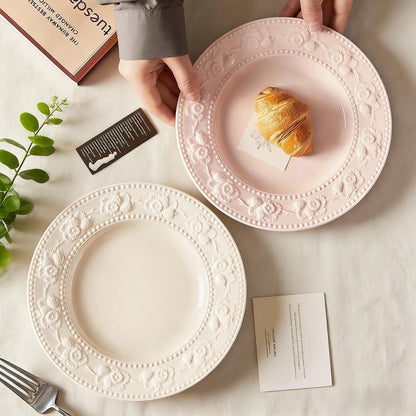 Two 8-inch ceramic plates with embossed floral borders in pastel pink and cream white, featuring a croissant on the pink plate.